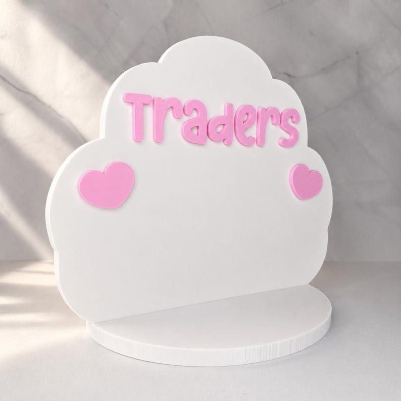 May include: A white cloud-shaped display stand featuring the pink text "Traders" in a rounded font, accented by two pink heart-shaped icons on either side. The display is mounted on a circular white base, providing a stable platform for items.