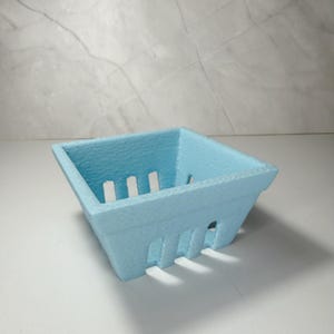 May include: A pale blue, square basket with vertical slots on the sides, resembling a berry punnet. The container has a textured finish and is empty. It is placed on a white surface. The dimensions are approximately 10 cm x 10 cm.