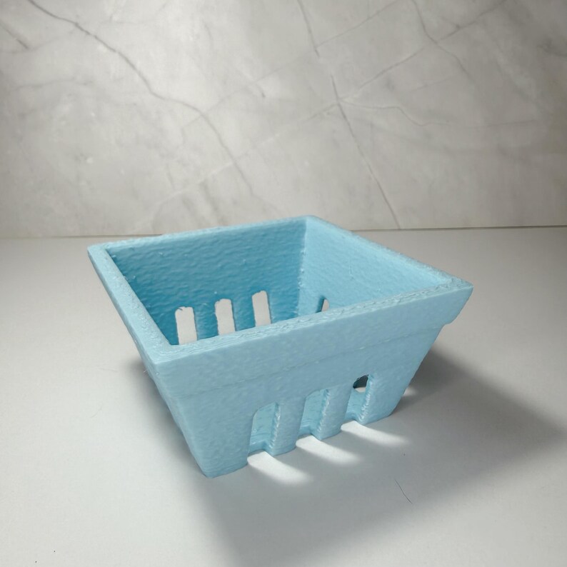 May include: A pale blue, square basket with vertical slots on the sides, resembling a berry punnet. The container has a textured finish and is empty. It is placed on a white surface. The dimensions are approximately 10 cm x 10 cm.