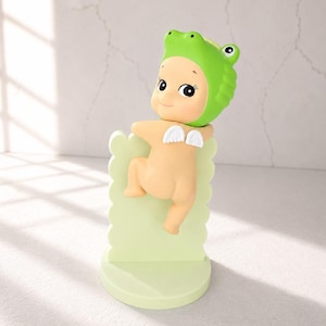 May include: A whimsical figurine of a nude baby wearing a green frog hat and small white wings. The figurine is perched on a light green scalloped stand with a round base. The baby has a smiling face and large, expressive eyes.