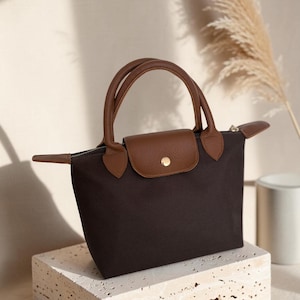 May include: A dark brown handbag featuring a trapezoidal silhouette, two short rounded handles, and a brown leather flap closure with a gold-tone snap button. The bag is constructed from a solid dark brown fabric with matching leather trim.
