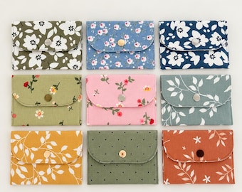 Puede incluir: Una colección de nueve estuches rectangulares de tela dispuestos en una cuadrícula, cada uno con un cierre de solapa con un botón a presión central. Los estuches muestran varios patrones que incluyen estampados florales, motivos de hojas y lunares en colores como oliva, azul, rosa, amarillo y óxido.