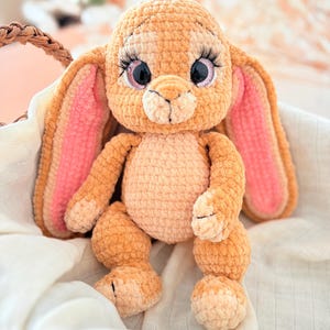 Può includere: Un coniglietto di peluche fatto a mano con lunghe orecchie, grandi occhi e lunghe ciglia. Il coniglietto è marrone chiaro con accenti rosa all'interno delle orecchie. È seduto in un cestino di vimini su un panno bianco.