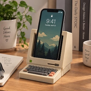 May include: A beige smartphone stand designed to resemble a vintage computer, featuring a small keyboard with grey and orange keys, a green power button, and a slot for a phone. The screen displays a mountain landscape with the time 9:41.