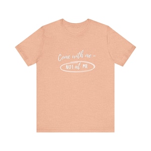 May include: A peach-colored t-shirt with the text "Come with me - NOT at ME" in white script and a white oval. The shirt has short sleeves and a crew neck.