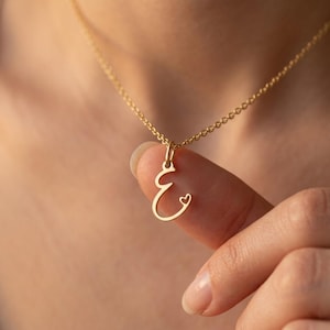 May include: A gold-colored necklace with a pendant shaped like the letter "E" with a small heart detail. The delicate chain and pendant are displayed against a blurred background, highlighting the jewelry's design.