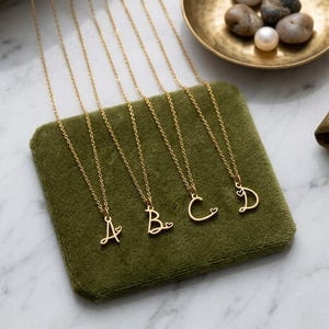 May include: Four gold-toned necklaces featuring cursive letter pendants A, B, C, and D. Each letter is crafted in a thin metal wire style and includes a small, integrated heart detail attached to the side of the character, suspended on delicate chains.