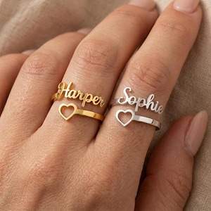 Può includere: Due anelli indossati su dita adiacenti. Un anello color oro presenta il nome Harper in corsivo sopra un piccolo intaglio a forma di cuore. Un anello color argento presenta il nome Sophie in corsivo sopra un piccolo cuore intagliato.