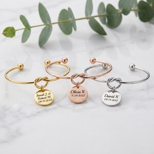 Op de afbeelding: Drie open armbanden in goud-, roségoud- en zilverkleurige tinten, elk met een centraal knoopontwerp en een ronde hanger. De hangers zijn gegraveerd met namen en datums: Sarah J. & Robert M. 06.12.2023, Olivia R. 11.24.2022 en David K. 02.14.2021.