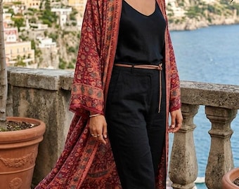 Op de afbeelding: Een lange, aan de voorzijde open kimono met een donkerrode en oranje bloemenprint, gedragen over een zwarte mouwloze top en een zwarte broek. De outfit is afgewerkt met een dunne bruine touwriem en bruine sandalen met bandjes.