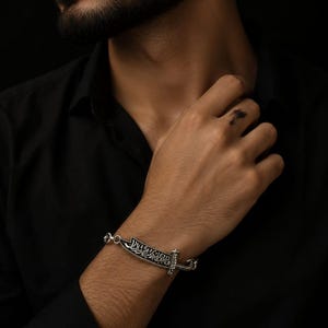 May include: Silver bracelet featuring a dagger design with intricate detailing and Arabic script. The bracelet is worn on a wrist against a black shirt. The dagger design is the focal point, showcasing the craftsmanship and cultural influence.