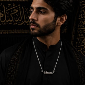 May include: A silver pendant necklace featuring a curved sword design. The necklace is worn against a black shirt. The background is a dark fabric with gold Arabic script.