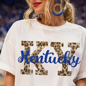 Peut inclure: Un t-shirt blanc à manches courtes arborant les lettres KY avec un motif léopard marron et noir, et le mot Kentucky écrit en écriture cursive bleue au centre. Le vêtement présente une encolure ronde et une coupe décontractée.