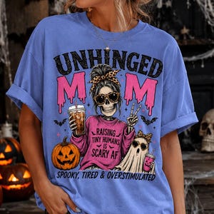 May include: A periwinkle blue t-shirt featuring a graphic print of a skeleton wearing a pink sweater, sunglasses, and a leopard print headband. The text reads UNHINGED MOM, RAISING TINY HUMANS IS SCARY AF, and SPOOKY, TIRED & OVERSTIMULATED.