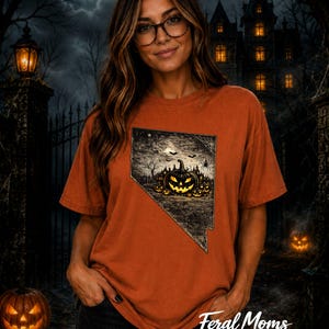 May include: A burnt orange short-sleeve t-shirt featuring a graphic print of the state of Nevada filled with a spooky Halloween scene. The design includes a glowing jack-o'-lantern, a haunted house, bats, and a spider web against a dark night sky.