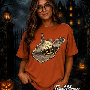 May include: A burnt orange short-sleeve t-shirt featuring a graphic of the state of West Virginia filled with a Halloween scene. The design includes a glowing jack-o'-lantern, a full moon, a haunted house, and a spider web against a dark background.