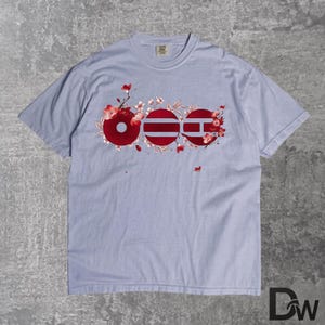 May include: A light blue t-shirt featuring a floral design with the word "OEE" in red, white, and blue. The design incorporates cherry blossoms and is centered on the front of the shirt. The shirt has a crew neck and short sleeves.