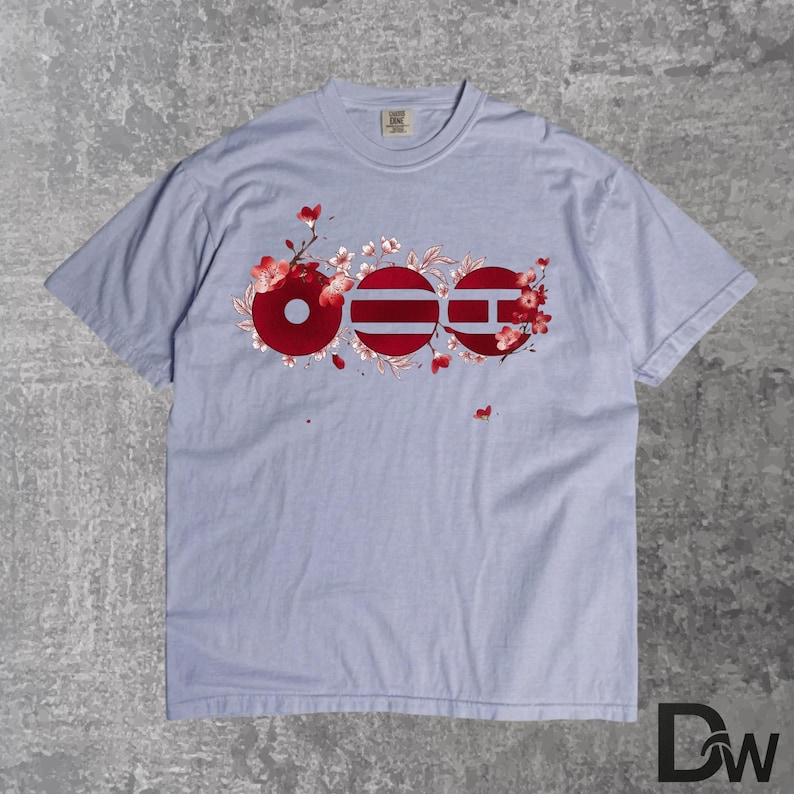 May include: A light blue t-shirt featuring a floral design with the word "OEE" in red, white, and blue. The design incorporates cherry blossoms and is centered on the front of the shirt. The shirt has a crew neck and short sleeves.