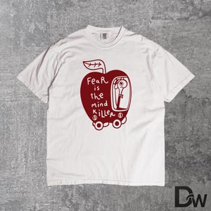 May include: White t-shirt featuring a red apple graphic with the text "Fear is the mind killer." The apple design includes a person inside a vehicle. The shirt has a crew neck and short sleeves.