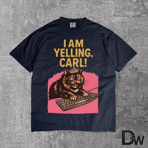 Pode incluir: Camiseta azul marinho com o texto "I AM YELLING, CARL!". O gr&aacute;fico apresenta um gato usando uma coroa, digitando em um teclado, em um fundo rosa. O gato &eacute; marrom e preto.
