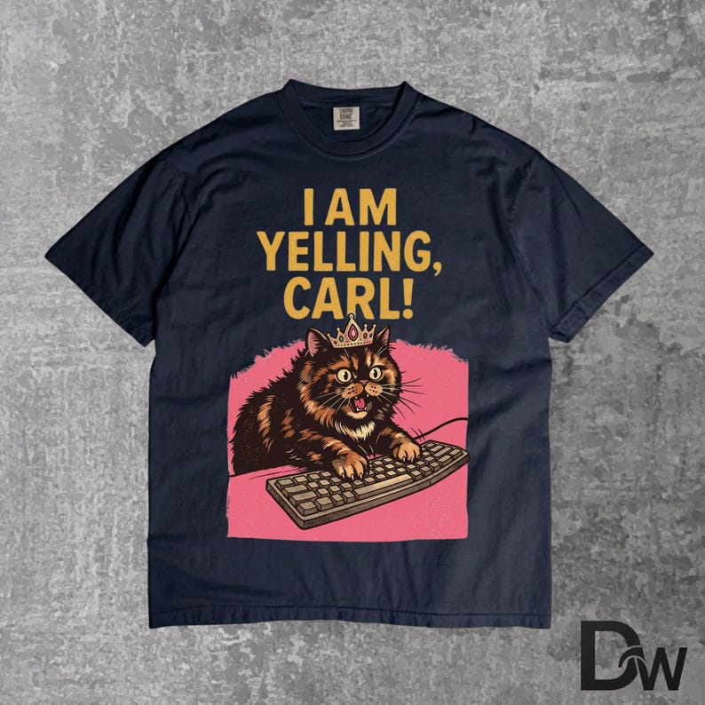 Pode incluir: Camiseta azul marinho com o texto "I AM YELLING, CARL!". O gr&aacute;fico apresenta um gato usando uma coroa, digitando em um teclado, em um fundo rosa. O gato &eacute; marrom e preto.