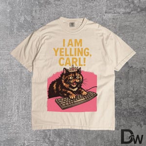 Pode incluir: Camiseta creme com um gato de desenho animado usando uma coroa e digitando em um teclado, em um fundo rosa. O texto "I AM YELLING, CARL!" &eacute; impresso acima do gato em amarelo. A camisa tem gola redonda e mangas curtas.