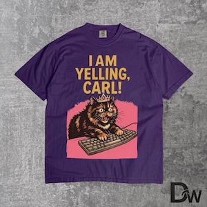 Pode incluir: Camiseta roxa com o texto "I AM YELLING, CARL!". O gr&aacute;fico apresenta um gato usando uma coroa, digitando em um teclado, em um fundo rosa. O gato &eacute; marrom e preto.