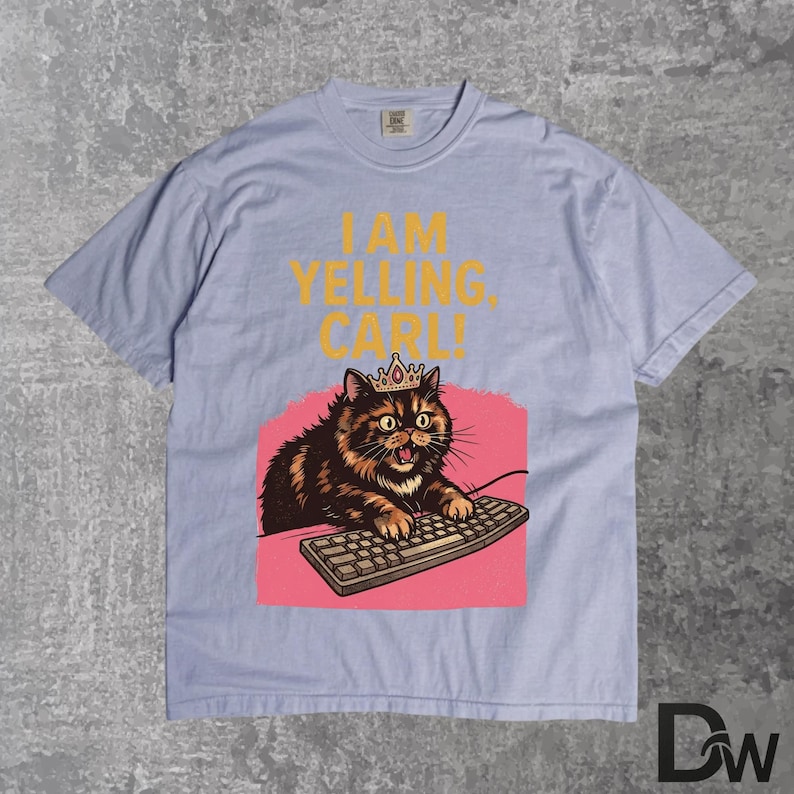 Pode incluir: Camiseta azul claro com um gato de desenho animado usando uma coroa e um teclado. O texto "I AM YELLING, CARL!" &eacute; impresso em amarelo acima do gato. O gato est&aacute; em um fundo rosa.