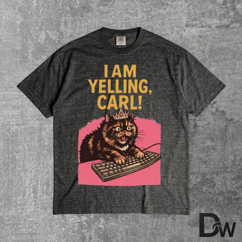 Pode incluir: T-shirt cinza escuro com o texto "I AM YELLING, CARL!". O gr&aacute;fico apresenta um gato usando uma coroa, digitando em um teclado, em um fundo rosa. O gato &eacute; marrom e preto.