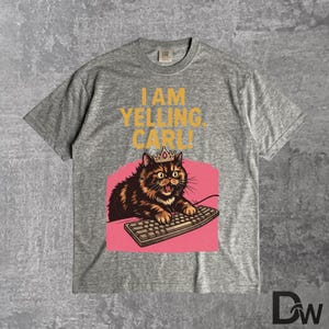 Pode incluir: Camiseta cinza com um gr&aacute;fico de um gato usando uma coroa e digitando em um teclado. O texto "I AM YELLING, CARLI" est&aacute; impresso acima da ilustra&ccedil;&atilde;o do gato. O gato &eacute; marrom e preto com fundo rosa.