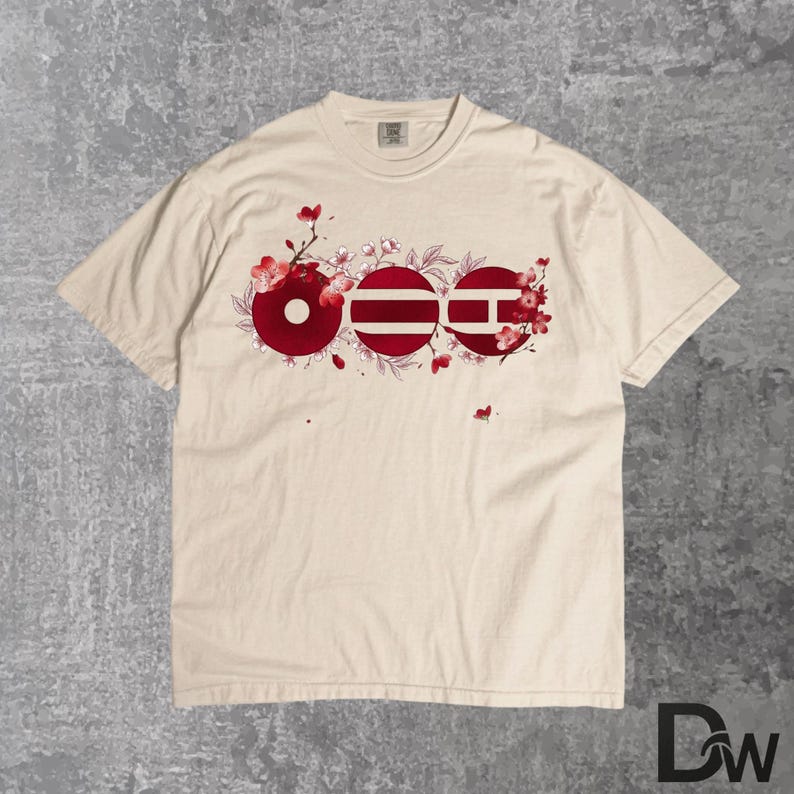 May include: Cream-colored t-shirt featuring a red floral design with the letters OEE. The design incorporates a circle, a striped circle, and a square with a horizontal line. The shirt has a crew neck and short sleeves.