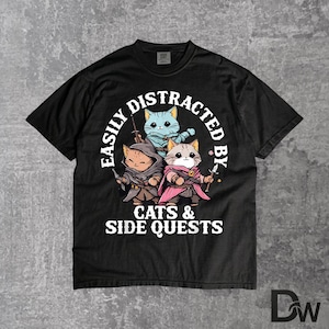 Peut inclure: T-shirt noir avec un motif de trois chats de dessin animé vêtus de costumes fantastiques. Le texte "EASILY DISTRACTED BY CATS & SIDE QUESTS" est en blanc. Les chats sont dans des tons orange, bleu et gris.
