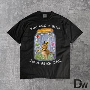 May include: A black short-sleeved t-shirt featuring a graphic of a cat wearing a bee costume inside a glass jar. The text reads YOU ARE A BUG IN A BUG JAR. The illustration includes a ladybird, dragonfly, butterfly, and bee surrounding the cat.