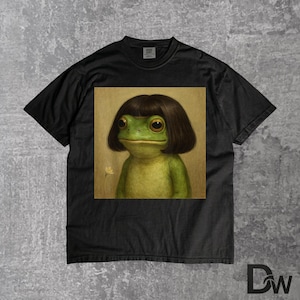 May include: Black t-shirt featuring a square image of a green frog with a brown bob haircut. The frog has large, expressive eyes and a small, white flower is visible in the background. The shirt is made of a soft material.