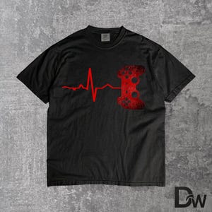 Puede incluir: Camiseta negra con un diseño de línea de latido cardíaco rojo que termina en un gráfico de mando de videojuego rojo. La camiseta tiene cuello redondo y mangas cortas. El diseño está impreso en la parte delantera.