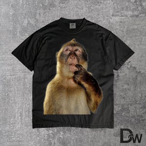 May include: Black t-shirt with a close-up image of a monkey, appearing to be deep in thought with its finger on its lips. The monkey has brown and tan fur. The t-shirt has a crew neck.