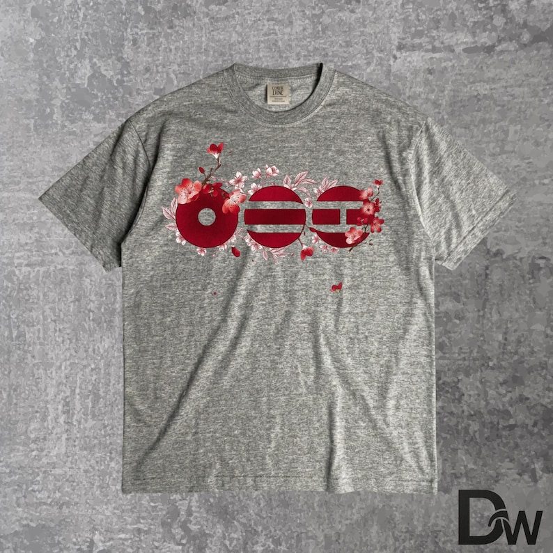 May include: A heather gray t-shirt featuring a floral design with the word "OEE" in red. The design incorporates cherry blossom branches and flowers. The shirt has a crew neck and short sleeves.