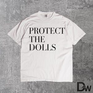 Puede incluir: Camiseta blanca con el texto "PROTECT THE DOLLS" en negro. La camiseta tiene cuello redondo y mangas cortas. La imagen está sobre un fondo gris texturizado.