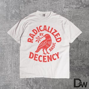 May include: A white t-shirt with a red graphic print. The design includes a bird holding a sprig, with the text "RADICALIZED BY BASIC DECENCY" in a distressed style. The shirt has a crew neck and short sleeves.