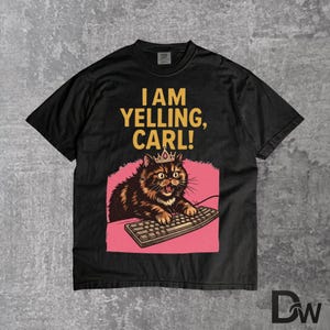 Pode incluir: Camiseta preta com um gato de desenho animado usando uma coroa e digitando em um teclado. O texto "I AM YELLING, CARL!" est&aacute; impresso acima do gato. O gato &eacute; marrom e preto com um fundo rosa.