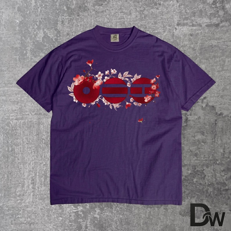 May include: A purple t-shirt featuring a floral design with the word "OOO" in red and white. The design incorporates cherry blossoms and a striped pattern within the letters. The shirt is made of a soft material.