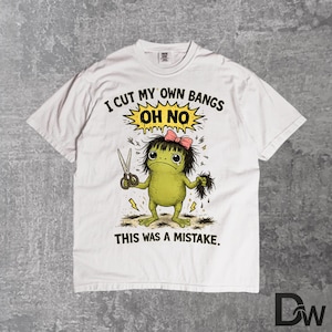Puede incluir: Camiseta blanca con un gráfico humorístico de una criatura verde cortándose el flequillo con unas tijeras. El texto dice: "I CUT MY OWN BANGS OH NO THIS WAS A MISTAKE". La criatura tiene un lazo rosa y una expresión de angustia.