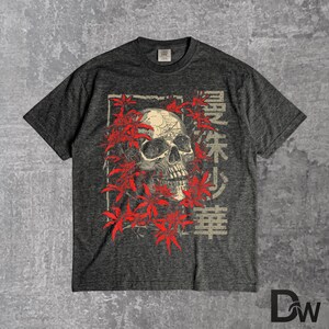 May include: Dark grey t-shirt with a skull graphic encircled by red leaves. Japanese characters are printed vertically on the right. The tee has a crew neck and short sleeves.