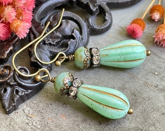 May include: A pair of green glass teardrop earrings with gold accents and clear rhinestone embellishments. The earrings are hanging from brass hooks.