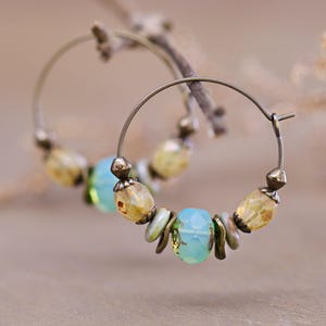 May include: A pair of hoop earrings featuring a thin bronze-toned metal wire. Each hoop is adorned with a sequence of beads, including faceted light blue glass beads, speckled yellow-toned beads, and small metallic spacers arranged symmetrically.