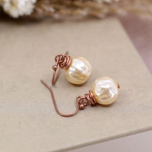 May include: A pair of pearl earrings with copper wire wrapping. The pearls are a cream color and the wire is a rose gold color.