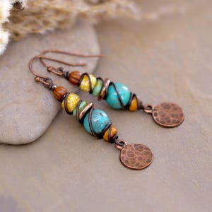 May include: Handmade copper wire earrings with turquoise, yellow, and brown beads. The earrings have a spiral design and a hammered copper disc at the bottom. The earrings are on a neutral background.