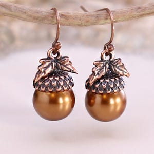 May include: A pair of drop earrings featuring a copper-toned metal acorn cap with two small leaves attached to the top. The base of each acorn is a smooth, round, metallic bronze-coloured bead. The earrings hang from simple copper-toned metal hooks.