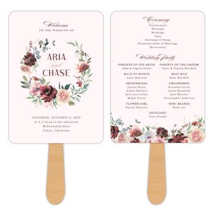 May include: Wedding fan featuring a floral wreath design with the names "Aria and Chase." The fan includes ceremony details and wedding party information. The handle is made of wood. The text "Reply by Design" is at the top left.
