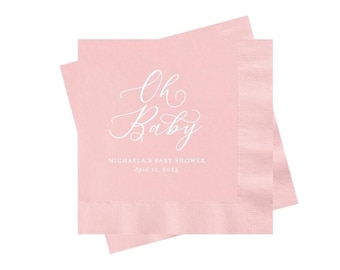 May include: Two square light pink paper napkins stacked at an angle. The top napkin features white text in a cursive script that reads Oh Baby, followed by the printed text MICHAELA'S BABY SHOWER and the date April 22, 2023 in a sans-serif font.